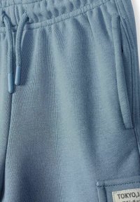 Pantalon de survêtement bleu clair avec taille élastique, cordon de serrage, poche latérale et patch rectangulaire cousu portant l'inscription "TOKYO.J".