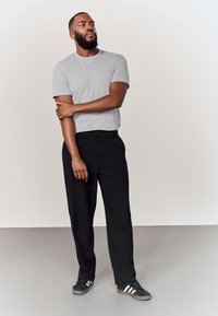 Harmaa lyhythihainen t-paita, mustat housut, seisoo vaaleanharmaalla lattialla. Mustat kengät, joissa valkoiset raidat. Neutraali tausta.