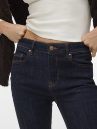 Person zieht dunkelblaue Jeans hoch, trägt ein weißes, geripptes Oberteil und eine braune, strukturierte Strickjacke.