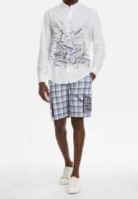 Muž v bílé košili s tlačítky a černým grafickým textem, v modrých kostkovaných krátkých kalhotách s bočními kapsami a v bílých teniskách, stojící s jednou rukou v kapse.