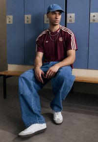 T-shirt adidas a righe bordeaux con logo rotondo, cappellino azzurro chiaro, jeans blu oversize e sneakers bianche con suola grigia.