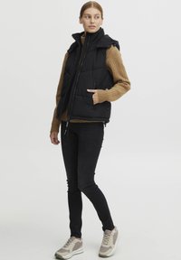 Svart vadderad väst med hög krage, dragkedja framtill och sidofickor. Parat med en beige räfflad tröja och svarta skinny jeans. Avslappnad skor.