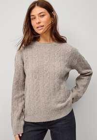 Jasnonude sweter w warkocze o miękkiej teksturze, z okrągłym dekoltem i długimi rękawami, w połączeniu z ciemnymi dżinsami.