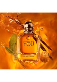 Klar glassparfymeflaske med et gyllent lokk, fylt med bernslig væske. Omgitt av grønne blader og brune vaniljestenger mot en oransje bakgrunn.