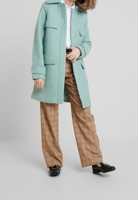 Cappotto in lana verde menta con un design strutturato, dotato di due tasche frontali e chiusura a bottoni. Abbinato a pantaloni larghi a quadri marroni e scarpe nere.