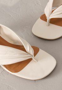 Weiße Leder-Sandalen mit flacher Sohle, die ein verdrehtes Überkreuz-Design auf dem Obermaterial und eine kontrastierende braune Lederinnensohle aufweisen.