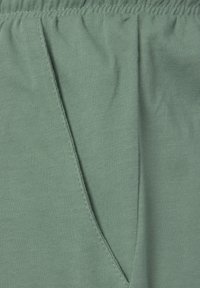 Zelená látka s jemnou žebrovanou texturou, s detaily boční kapsy a elastickým pasem. Hladký povrch a lehký materiál.