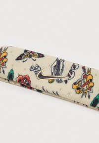 Turbante de malha em creme com padrões florais em vermelho, verde e amarelo. Apresenta um detalhe do logotipo da Nike em dourado bordado. Textura suave.