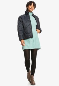 Black quilted puffer jacket with a stand collar, worn over a mint green ribbed dress, paired with black tights and brown ankle boots.