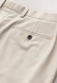 Pantalon beige habillé montrant la poche arrière avec un bouton noir et une boucle de ceinture, mettant en valeur la texture du tissu et les détails des coutures.