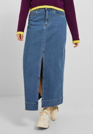 Persoon draagt een blauwe denim midi-rok met een split aan de voorkant, gecombineerd met beige sneakers en een paarse trui met gele boorden en zoom.