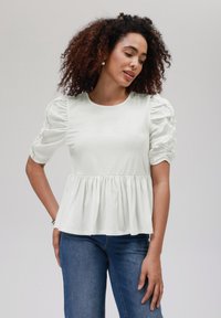 Light beige puff-sleeve top with gathered shoulders and a flared hem, made of soft, smooth fabric, paired with blue jeans.