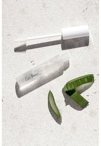 Klar tube med svart skriptemblem, hvit kork og applikatorbørste; to grønne aloe vera-blader, teksturert overflate, lys bakgrunn.