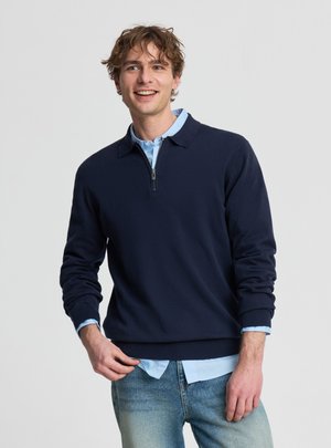 Giovane uomo sorridente, indossa un maglione blu navy con zip al quarto sopra una camicia azzurra e jeans chiari, in piedi davanti a uno sfondo semplice.