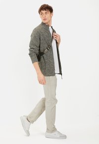 Grauer gestrickter Cardigan mit hohem Kragen, getragen über einem weißen Hemd, kombiniert mit hellbeigen Hosen und weißen Turnschuhen. Verstellbares schwarzes Riemendetail.