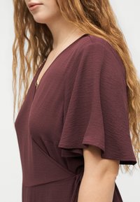 Γυναίκα με μακριά κυματιστά μαλλιά που φοράει ένα μακρύ φόρεμα μπορντό με V-λαιμόκοψη και φτερατά μανίκια, εμφανιζόμενη από τους ώμους μέχρι τη μέση.