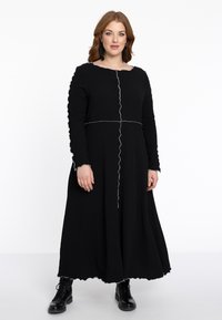 Schwarzes langärmliges Kleid mit Textur, gewelltem Saum und weißen Nähdetails. Kombiniert mit schwarzen Schnürstiefeln.