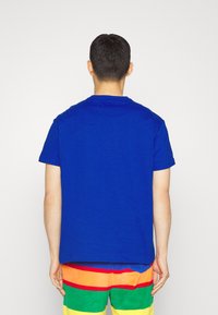 En person med kort mörkt hår som bär en enfärgad blå t-shirt och randiga shorts i regnbågens färger, stående vänd bort från kameran mot en helt vit bakgrund.