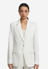 Vit blazer med strukturerat mönster, enfaldig knäppning, slagkrage och två framfickor. Ärmarnas längd är lång med knappar vid ärmsluten.