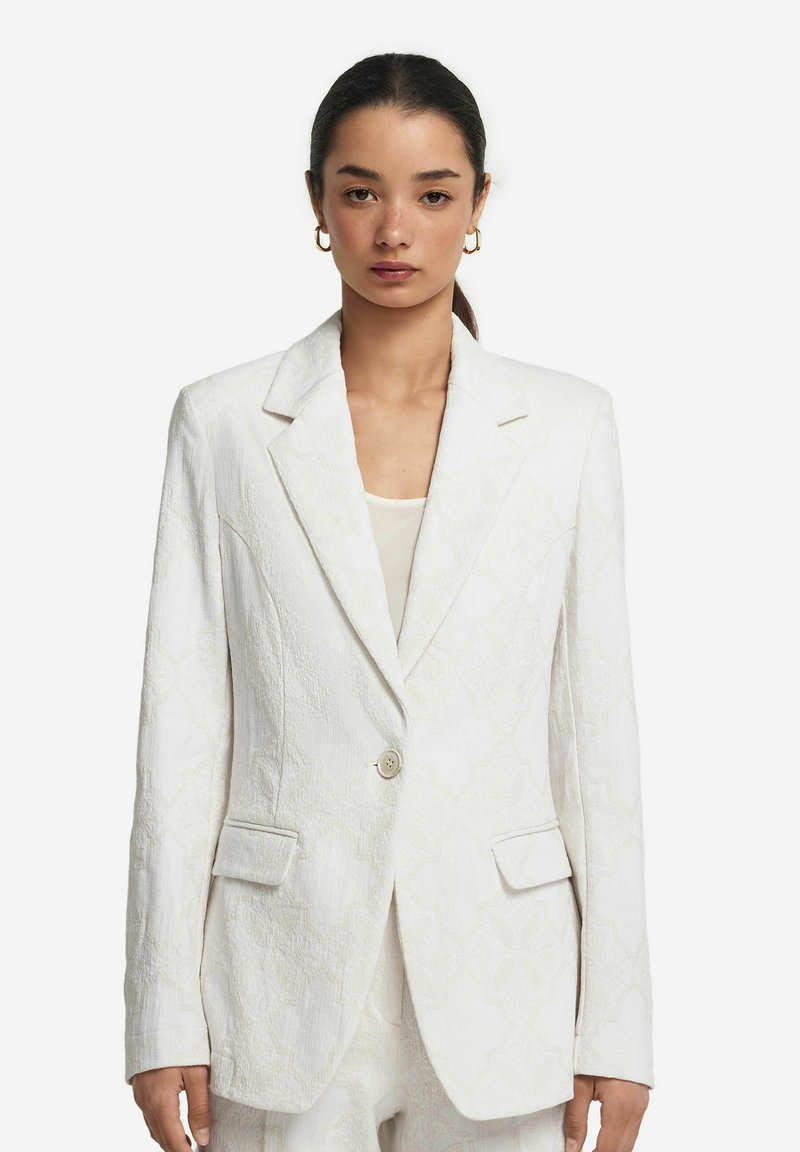 Vit blazer med strukturerat mönster, enfaldig knäppning, slagkrage och två framfickor. Ärmarnas längd är lång med knappar vid ärmsluten.