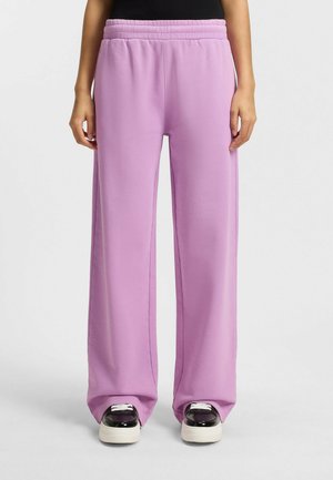 Osoba nosząca luźne liliowe spodnie dresowe z elastycznym pasem oraz czarno-białe trampki na jednolitym białym tle.
