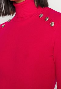 Gros plan d'une personne portant un pull à col roulé côtelé rose vif avec trois boutons décoratifs dorés sur chaque épaule.