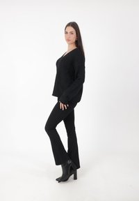 Schwarzer, übergroßer Pullover mit V-Ausschnitt, kombiniert mit schwarzen, ausgestellten Jeans und spitzen schwarzen Absatzstiefeln, vor einem schlichten weißen Hintergrund präsentiert.