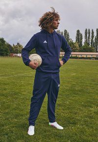 Marineblå treningsdrakt laget av mykt stoff, med hettegenser og tettsittende bukser. Spilleren holder en teksturert fotball, iført hvite fotballsko med fargerike detaljer.