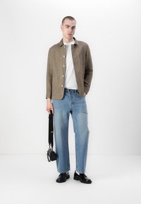 Tan linen blazer with large buttons, white shirt, blue relaxed-fit jeans, and black shoes. Holds a small black bag with a shoulder strap.