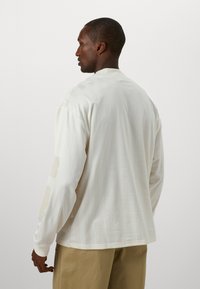Camicia a maniche lunghe in tessuto avorio con una texture liscia. Presenta sottili righe verticali e grafiche beige sulle maniche. Vestibilità casual.