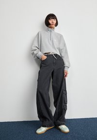 Svijetlosivi dukserica s patentnim zatvaračem na ovratniku, uparen s lepršavim tamnosivim cargo hlačama s bočnim džepovima. Pojedinac nosi patike u mint zelenoj boji.