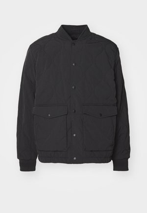 Black quilted jacket with a stand collar, front snap closure, and two large flap pockets. Features ribbed cuffs and textured fabric.