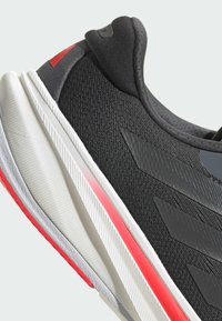 Black athletic shoe with textured mesh upper, gray accents, white midsole, and red detailing on the heel and sole, featuring a sleek design.