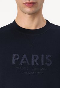 Felpa blu navy con scollo a girocollo. Il design del testo presenta "PARIS" in evidenza, con testo più piccolo sottostante. Texture morbida.