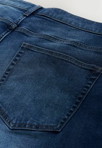 Tmavomodré denimové džínsy so zadnou vreckom s jemnou štruktúrou a kontrastným prešívaním. Sú viditeľné čisté línie a klasické dizajnové detaily.