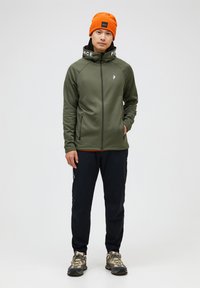 Grøn zip-jakke med høj krave og teksten "PEACE", orange hue, mørke bukser og vandrestøvler; struktureret materiale, atletisk pasform.