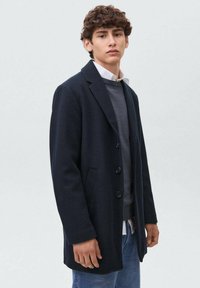 Joven vestido con un abrigo azul marino sobre un suéter gris y una camisa blanca, de pie frente a un fondo gris claro liso.