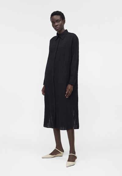 Γυναίκα στέκεται φορώντας μακρύ μαύρο φόρεμα με κουμπιά και γιακά και κρεμ μυτερές μπαλαρίνες με λουράκια σε λευκό φόντο.