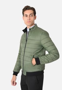 Giacca bomber imbottita verde con colletto alto, chiusura con zip, tasche laterali e polsini e orlo a coste neri. Materiale liscio e leggero.