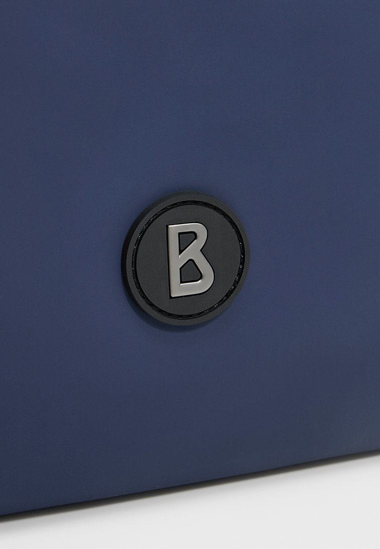 Granatowa torba o gładkiej teksturze, z okrągłym czarnym akcentem logo z srebrnym "B" na środku.