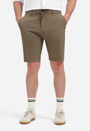 Mann trägt olivgrüne maßgeschneiderte Shorts mit seitlichen Taschen, weiße Socken mit blauen Streifen und weiße Schnürschuhe, steht mit den Händen in den Taschen.