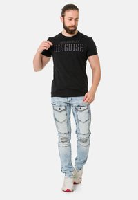 Schwarzes T-Shirt mit dem Aufdruck "CIPO AND BAXX DISGUISE", kombiniert mit hellblauen, stark verwaschenen Jeans mit Taschen und Verzierungen. Weiße Sneakers.