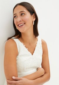 Abito bianco in pizzo con scollatura a V e motivi floreali. Il modello ha lunghi capelli scuri e indossa orecchini a cerchio in argento.