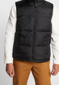 Man som bär en svart pufferväst med frontdragkedja över en vit långärmad skjorta och bruna byxor, med avslappnade händer vid sidorna.