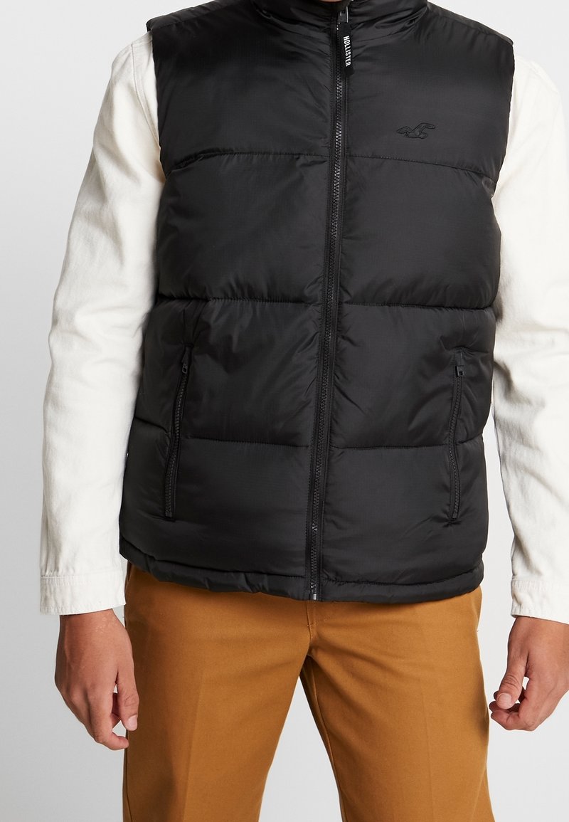 Man som bär en svart pufferväst med frontdragkedja över en vit långärmad skjorta och bruna byxor, med avslappnade händer vid sidorna.