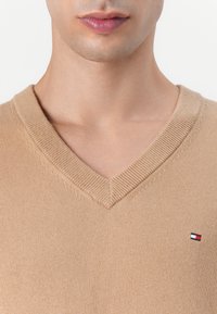 Beige V-hals trui van breiwerk, met een klein, merklogo aan de linkerzijde. Soepele textuur en aansluitend ontwerp.
