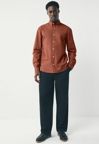 Camicia a maniche lunghe di colore ruggine con colletto button-down, abbinata a pantaloni larghi blu navy e scarpe nere con suola in legno.