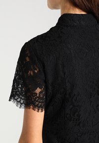 Black lace top featuring a high collar and short fringed sleeves, with floral patterns throughout the fabric and a textured design.