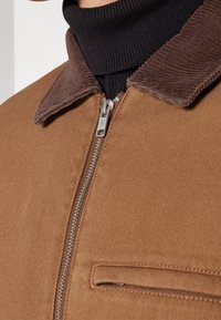 Brun fløjlsk jacket med lynlås lukning, der har en mørkebrun fløjlsk krave og en brystlomme. Ribbede sorte turtleneck under.