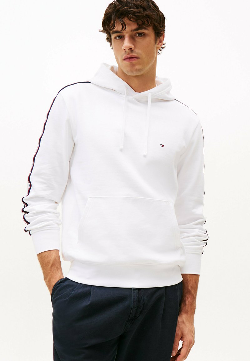 Sudadera blanca con capucha y bolsillo canguro, con franjas negras y rojas a lo largo de las mangas. Fabricada en tela suave, de corte casual.
